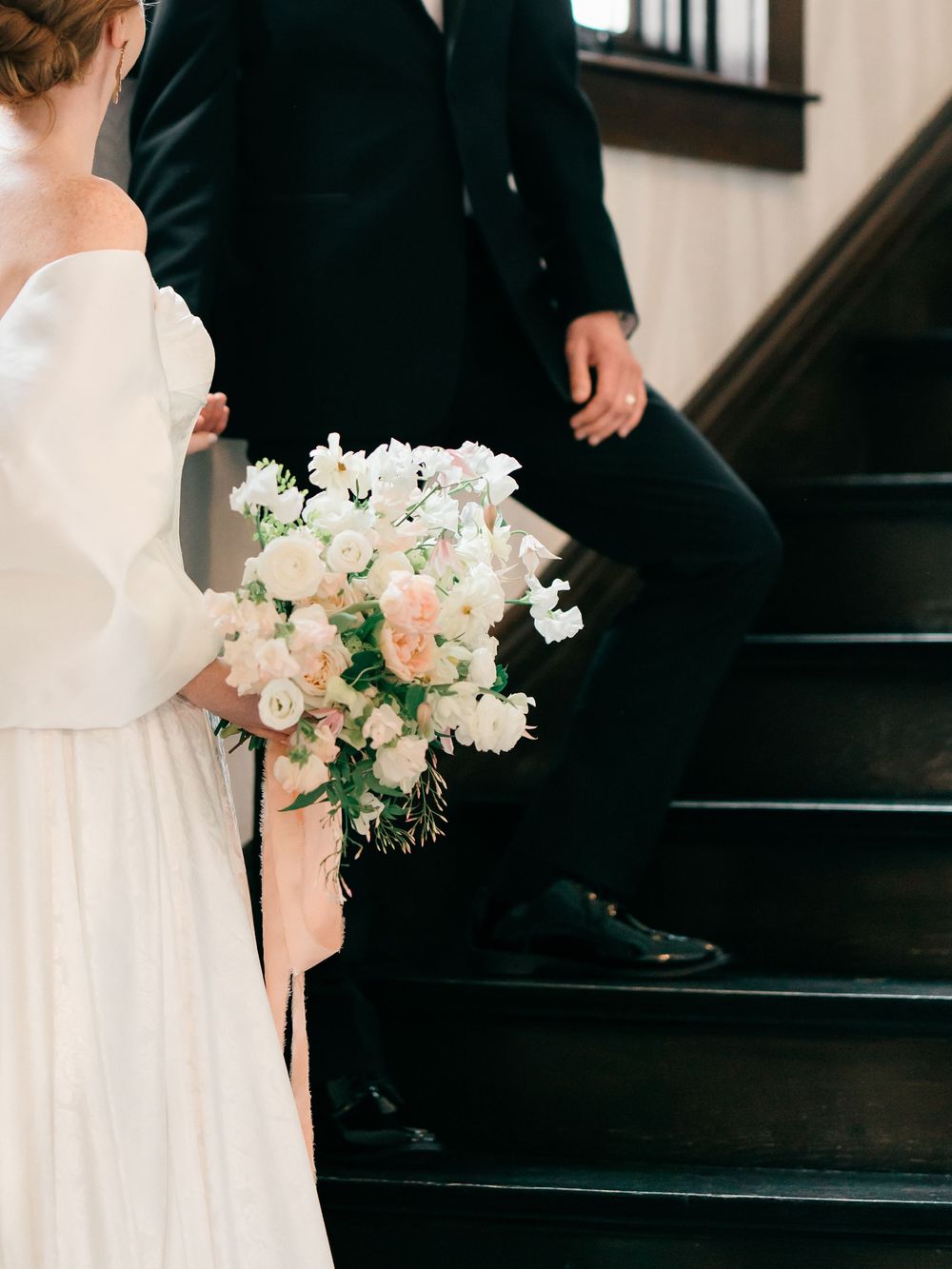 A black tuxedo and white wedding dress are captured in a romantic moment on dark wooden stairs with a blush bouquet.