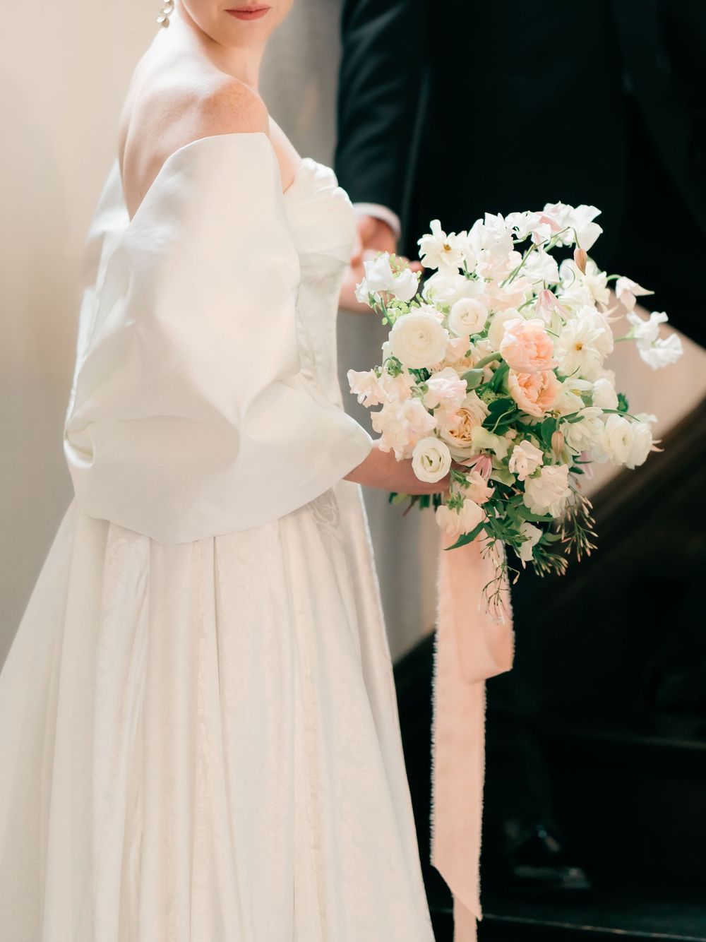 White wedding bouquet with delicate pink and peach roses and greenery against an ivory wedding dress.