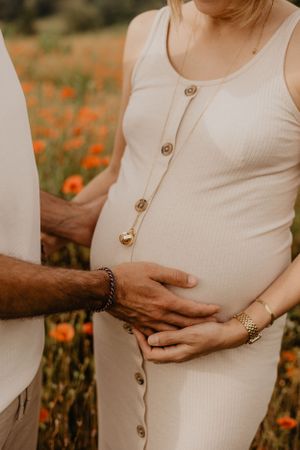 Close-up of a pregnant belly in a cream-colored button-front dress with loving hands embracing.