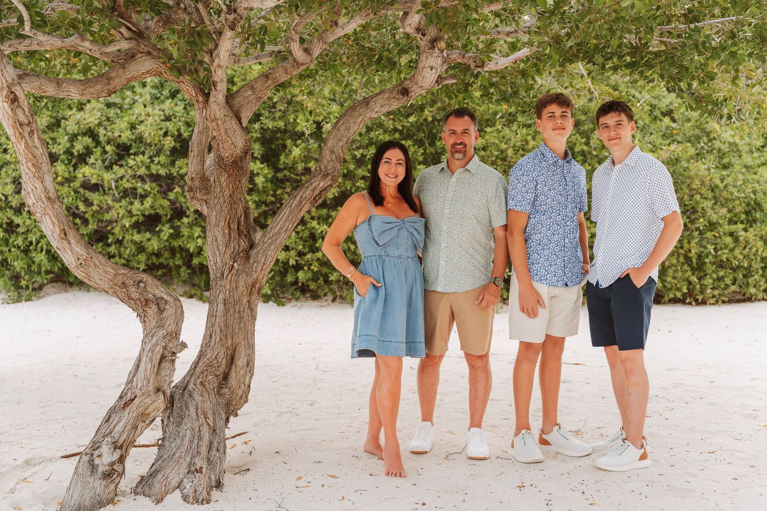 A group in coordinated blue and white outfits poses on a sandy beach near a twisted tree trunk.