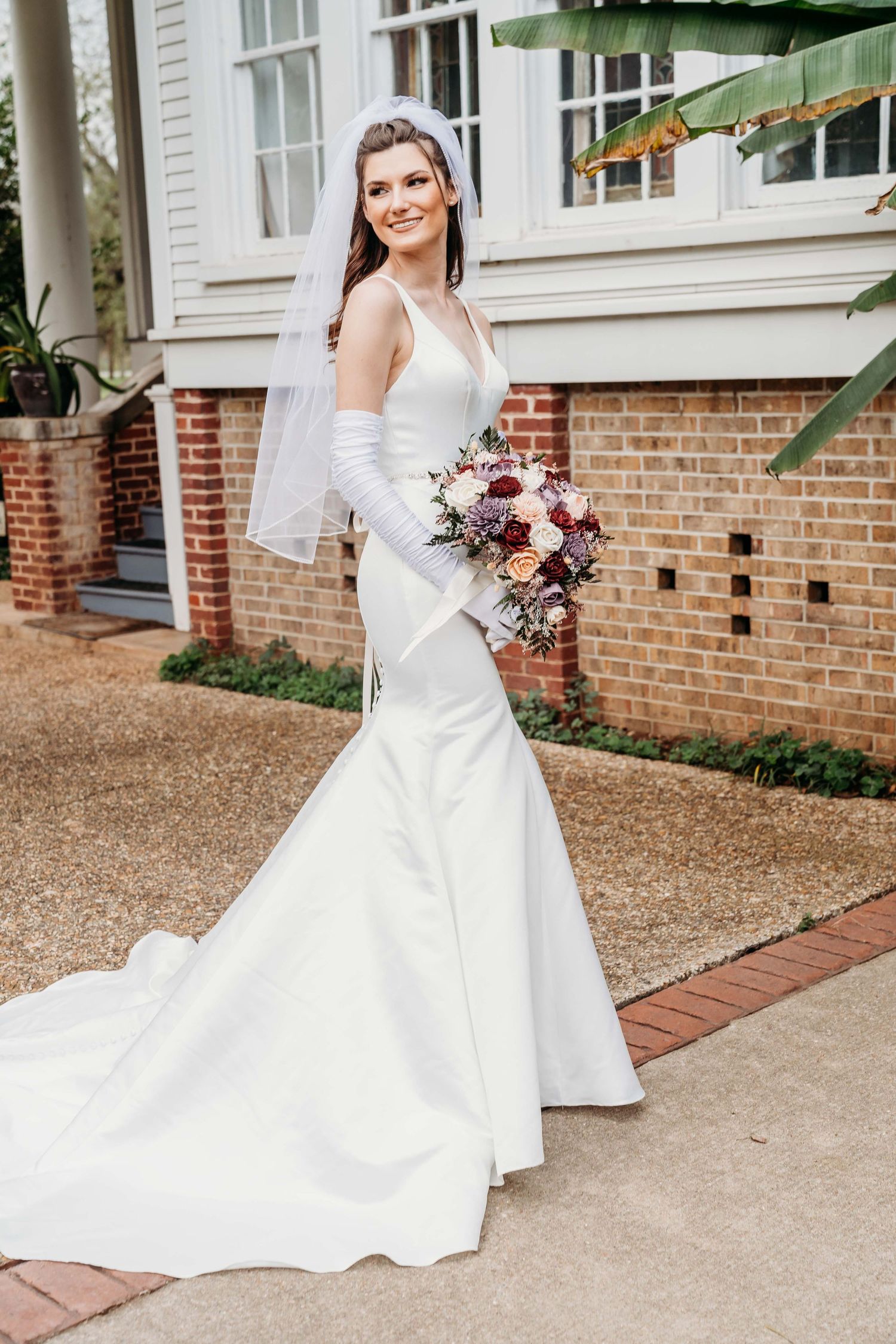 Elegant white mermaid wedding gown with long train and pink floral bouquet photographed outside brick building.