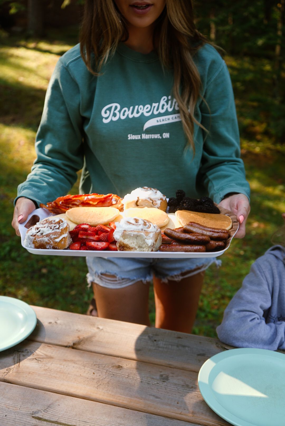 Person holding a tray of food while standing at an outdoor wooden table.