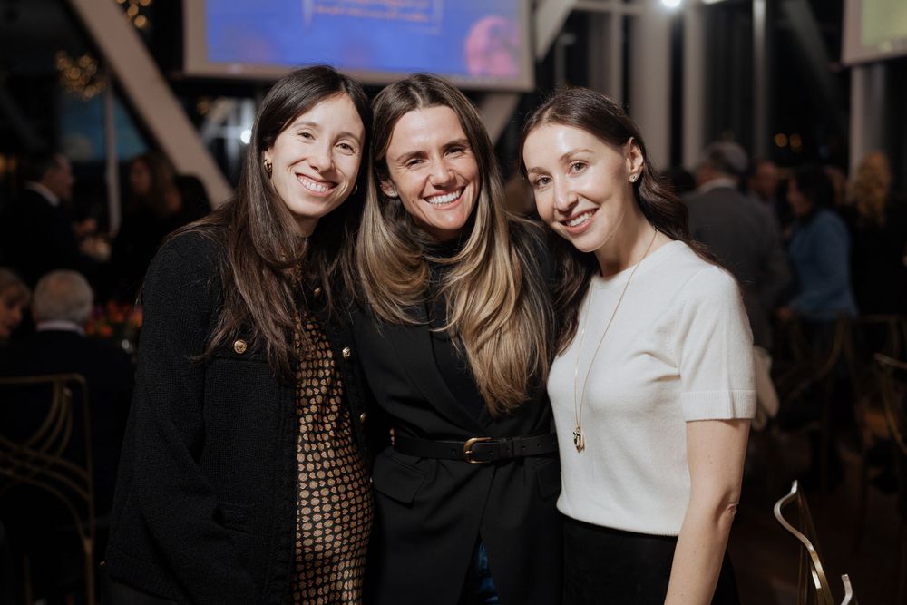 Three friends smiling together at a social gathering with dim lighting and crowd in background.