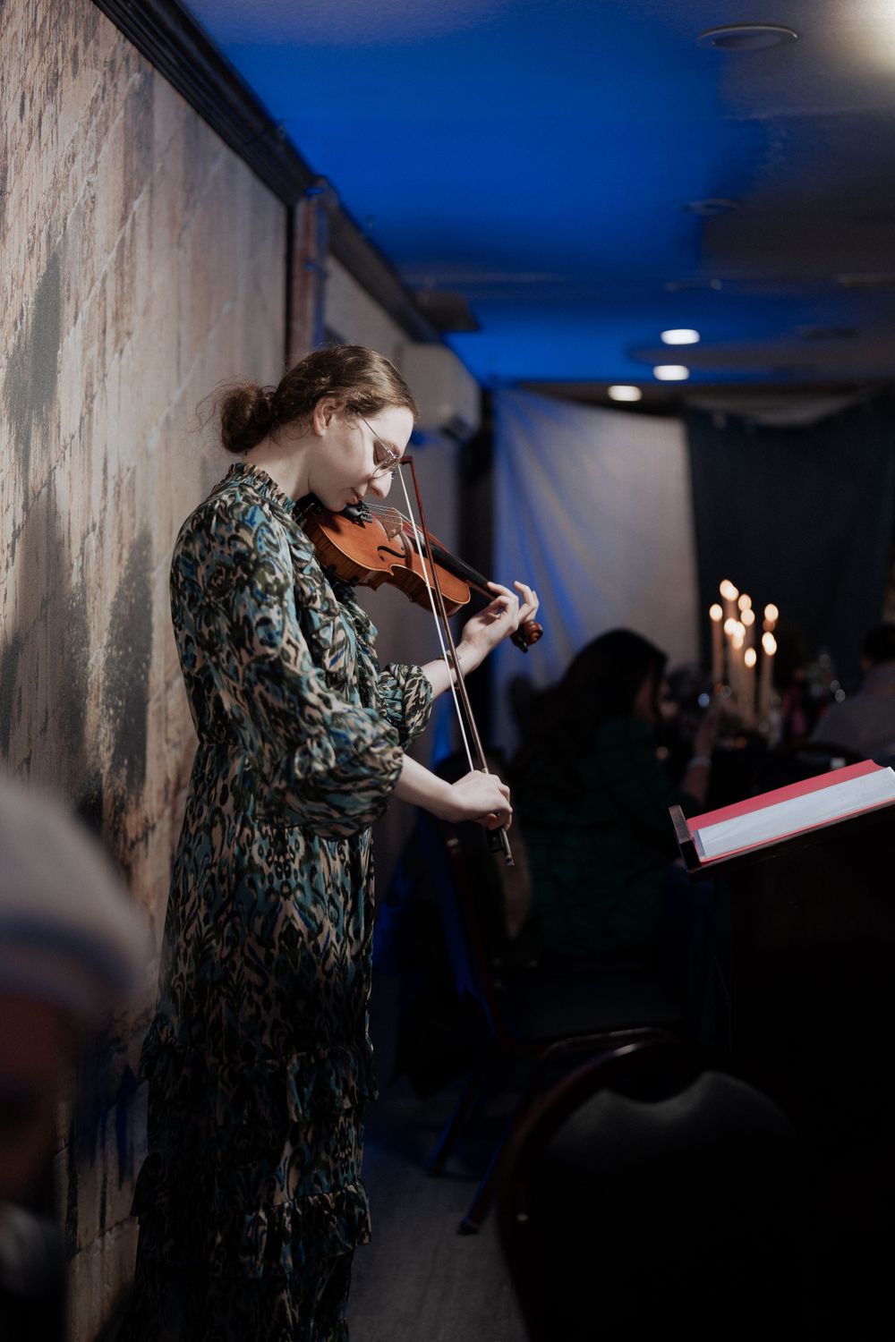 Violinist performing in patterned dress against blue-lit background with candles visible.
