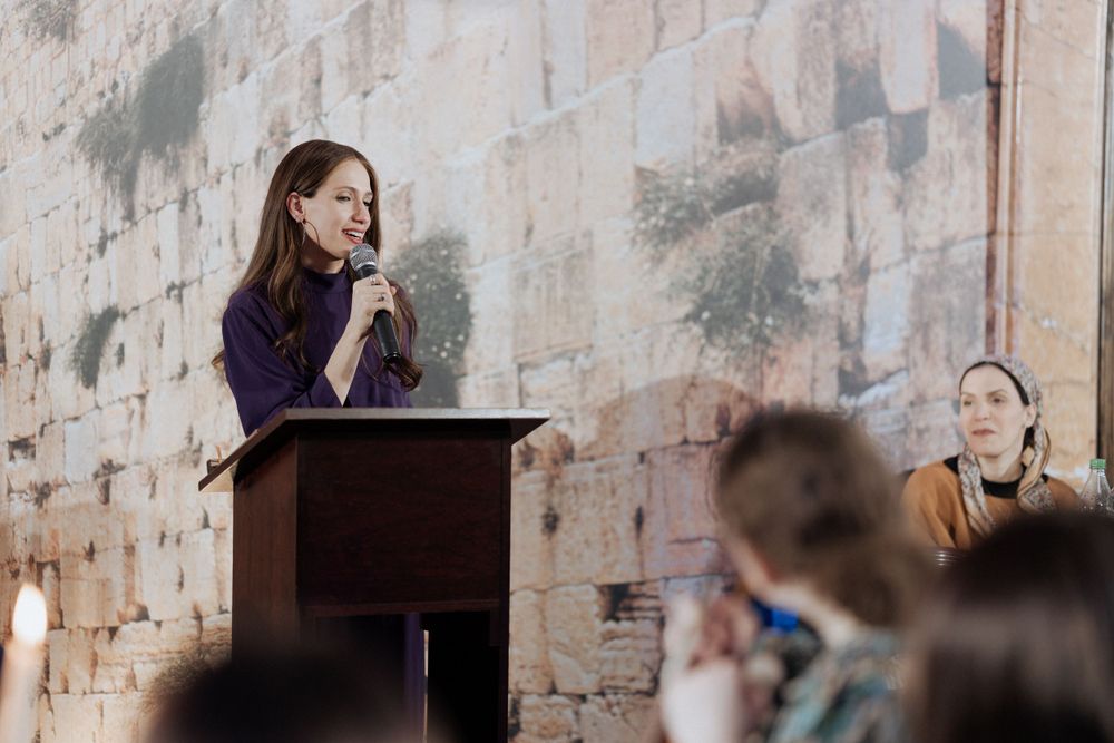 A speaker at a wooden podium addresses an audience against a stone wall background.