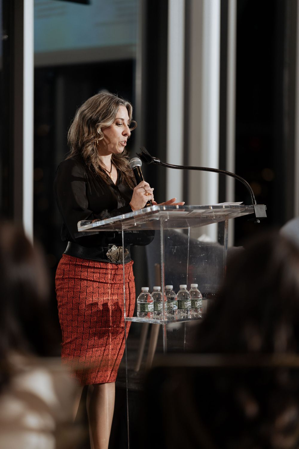 A speaker in business attire presenting at a clear podium with water bottles.