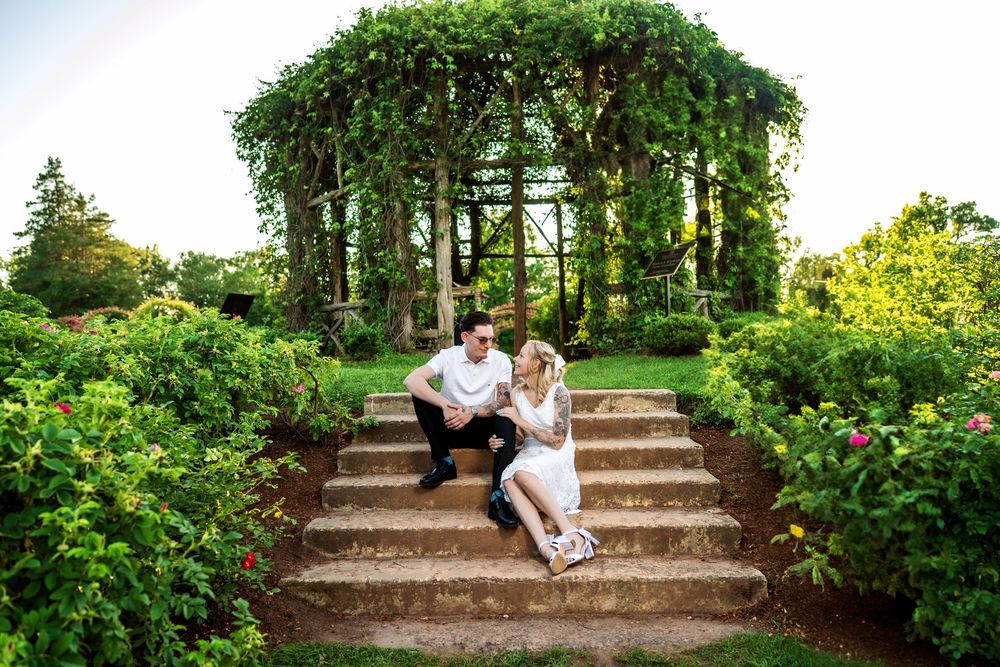 A couple sits on garden steps beneath a lush green pergola covered in vines.