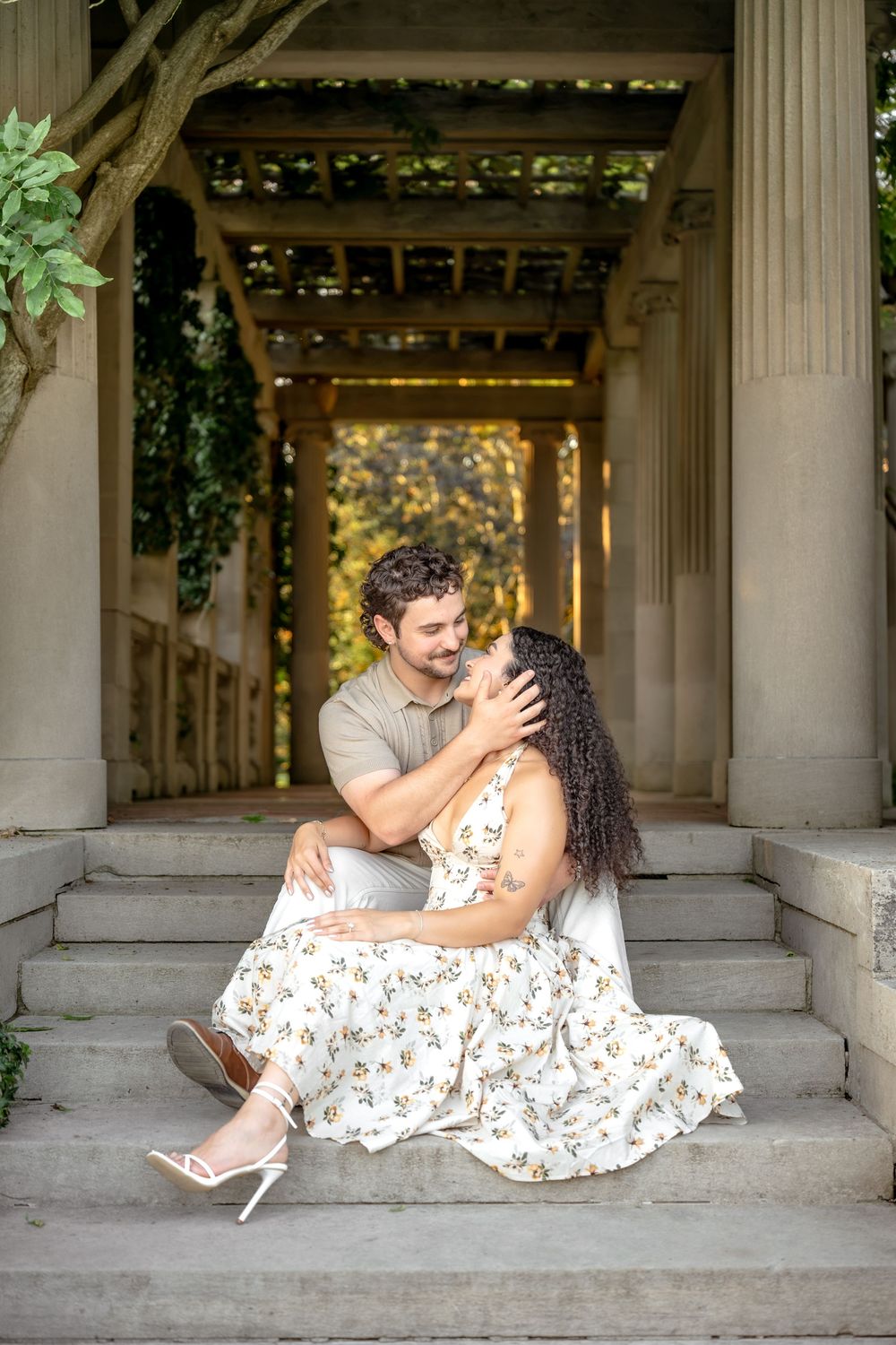 Couple sharing a romantic moment on stone steps under a pergola, woman in floral dress, man in gray shirt.