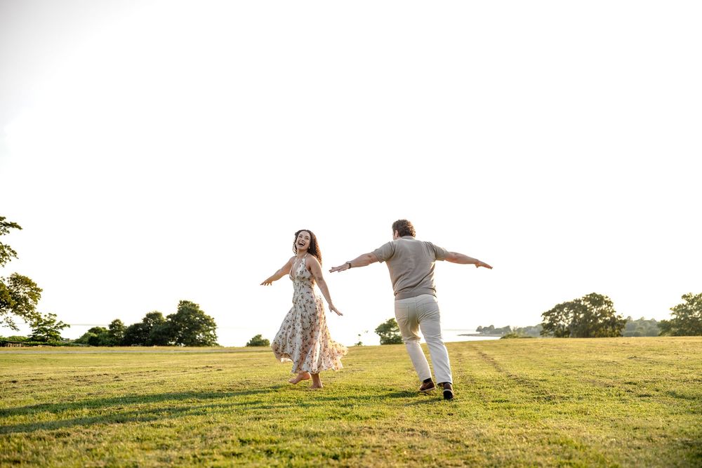 Couple dancing joyfully in a sunlit green field surrounded by trees during golden hour.