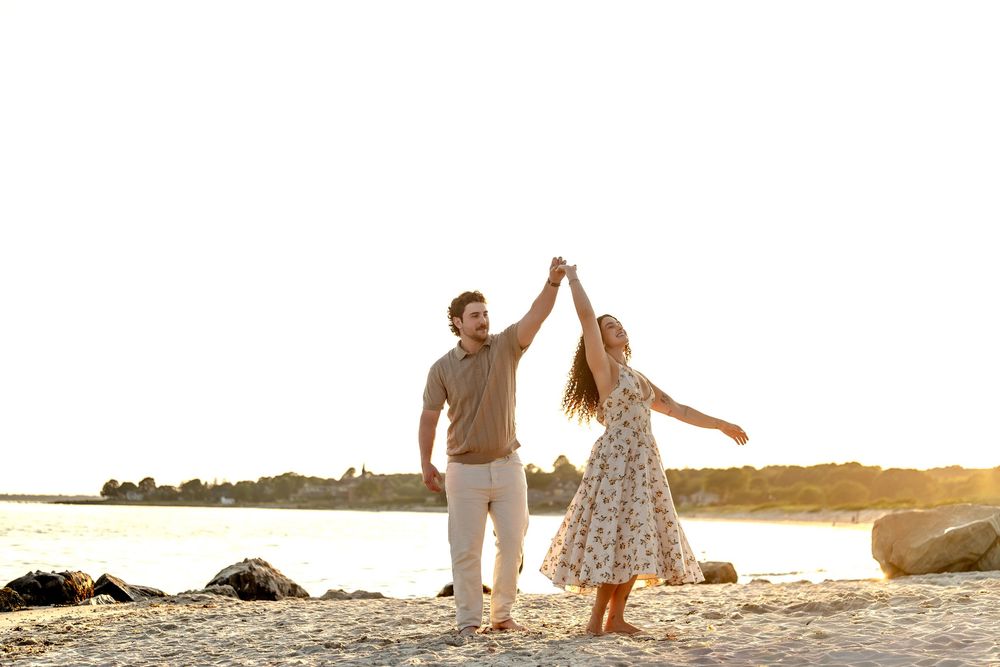 Couple dancing joyfully on a sunny beach at golden hour, holding hands with waves and rocks in background.