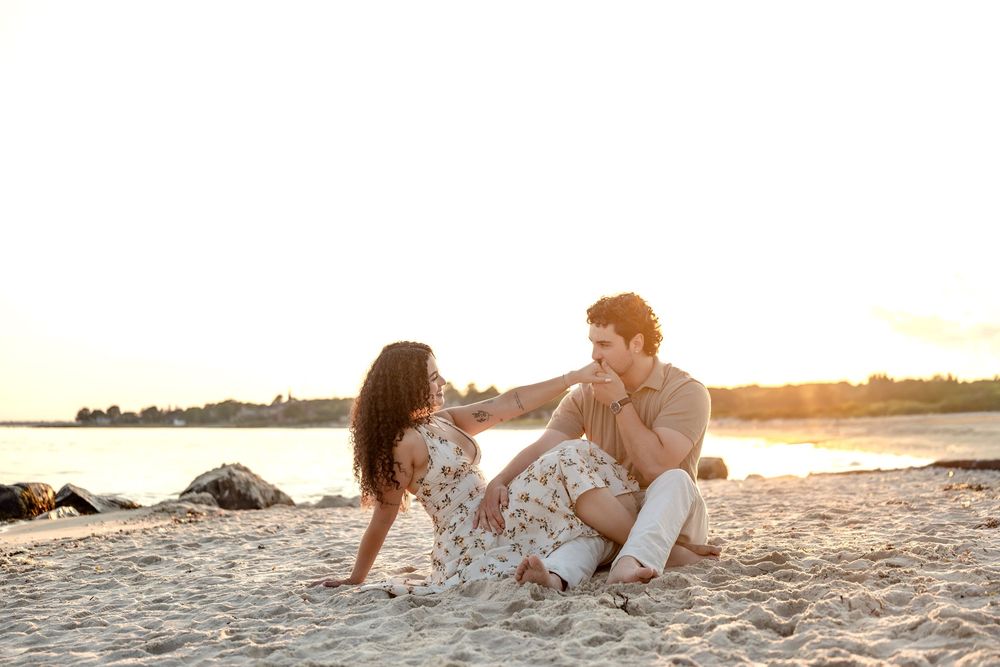 Couple sitting on sandy beach at sunset, sharing a romantic moment with golden light reflecting on the water.