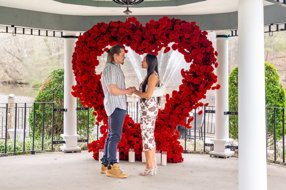 Couple holding hands in front of a large red rose heart arch in a white gazebo.