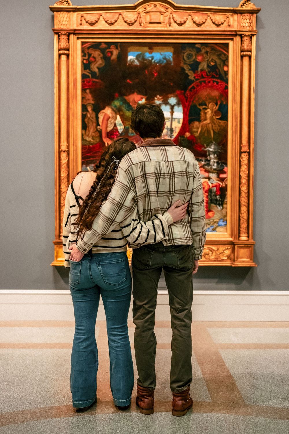 Two people viewing framed artwork in a museum gallery with ornate gold frames.