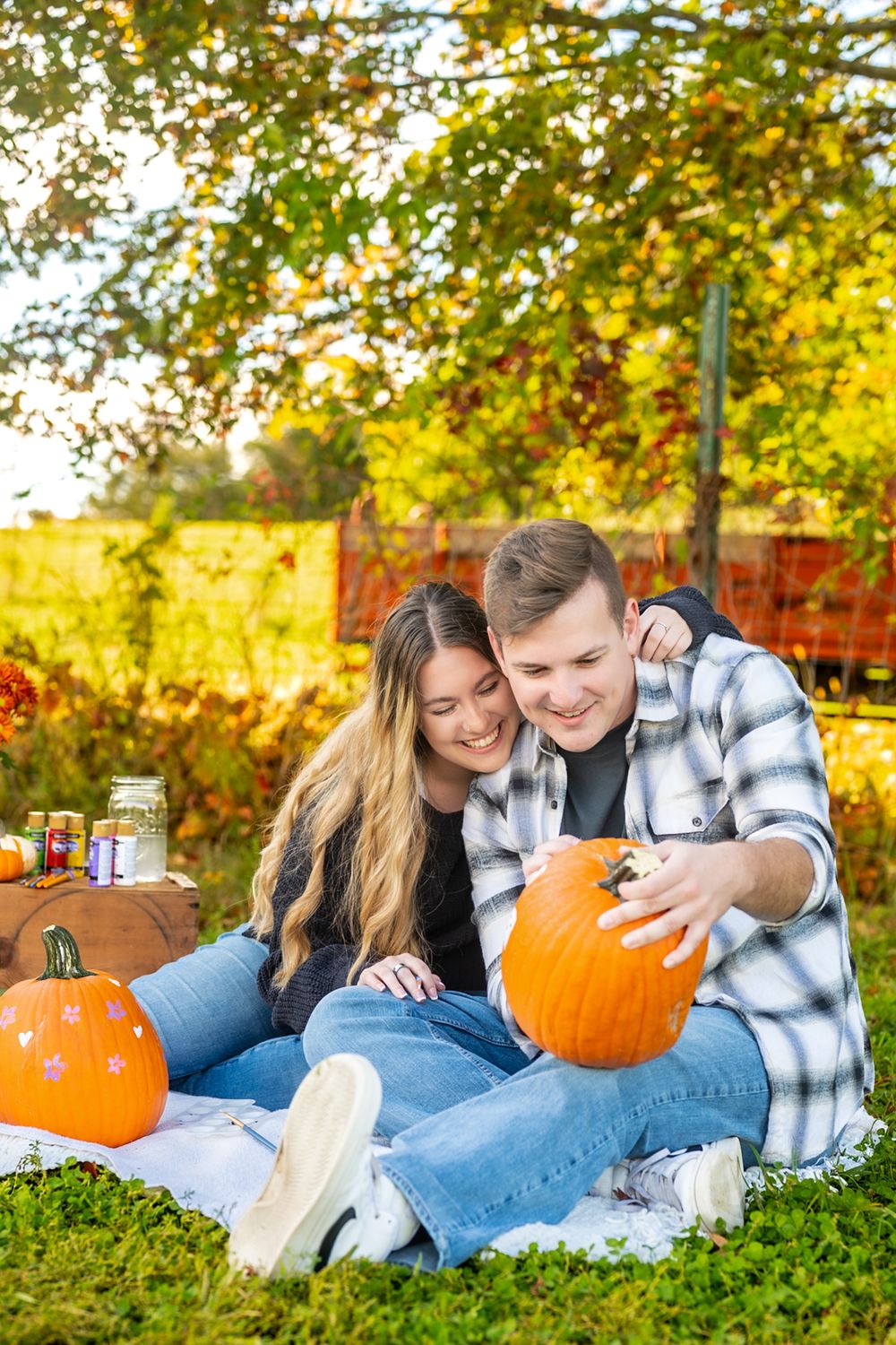 A couple sits on a blanket with pumpkins during a cozy autumn photoshoot in a park.