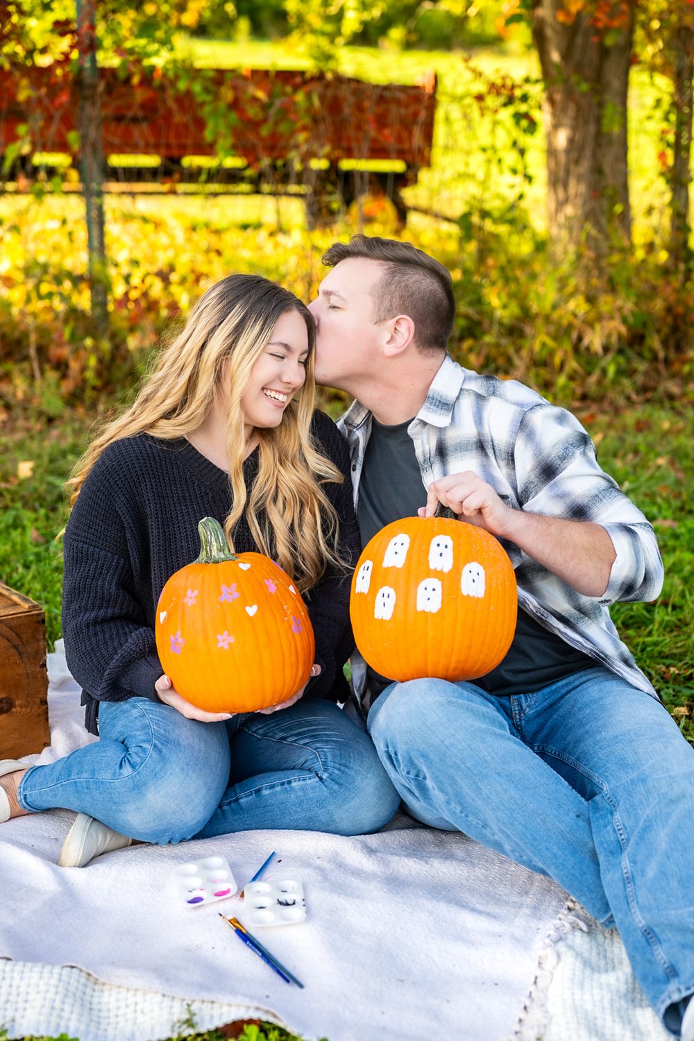 Two people share a romantic moment while decorating pumpkins on a blanket in fall foliage.