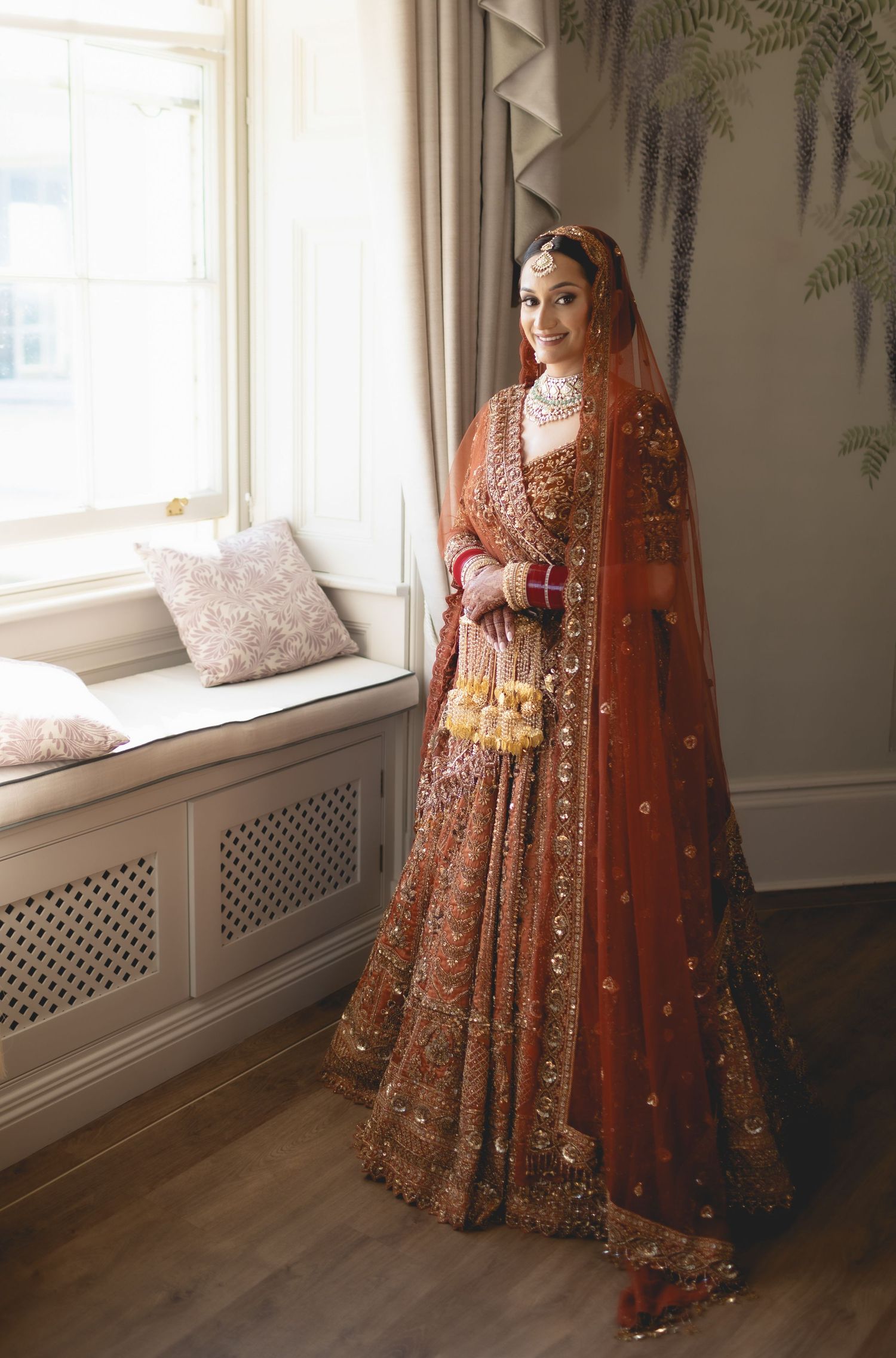 South Asian bride in an ornate rust-colored lehenga with intricate embroidery poses elegantly by a window.