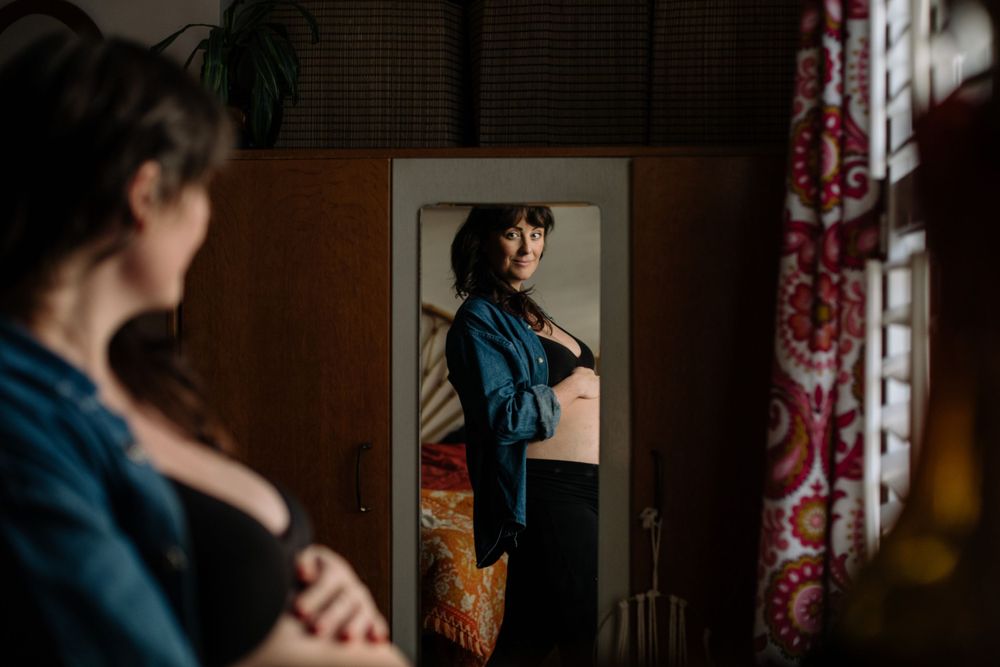 A person in a teal sweater looks at their reflection in a mirror with red curtains visible in the background.