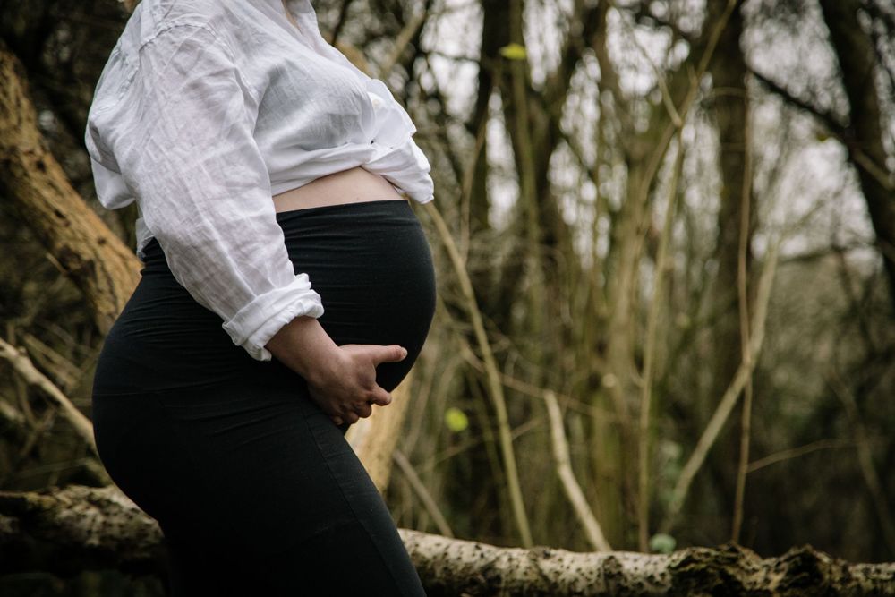 Side view of pregnant woman in white shirt and black pants standing by tree in moody forest setting.