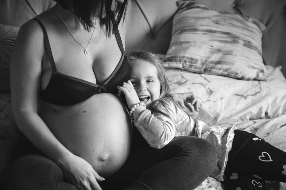 A black and white photo of an intimate maternity moment showing a pregnant belly with a young child resting nearby on a bed.
