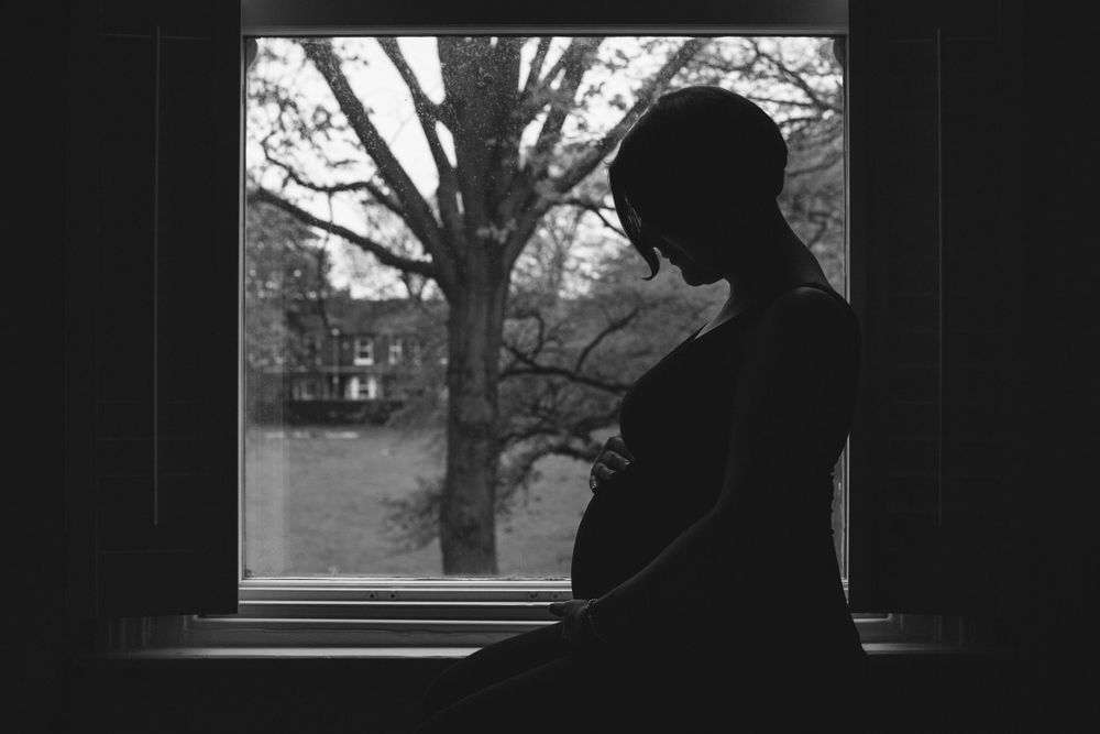 A silhouetted figure gazes contemplatively through a window at a tree and buildings in black and white.