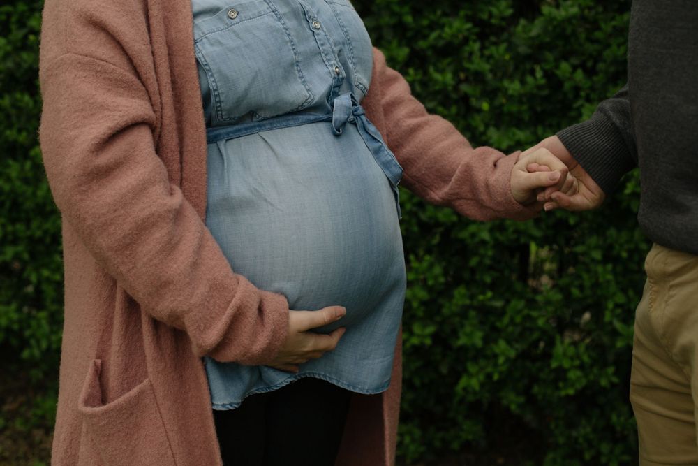 A pregnant person in a denim dress and brown cardigan holds hands with someone while cradling their belly against greenery.