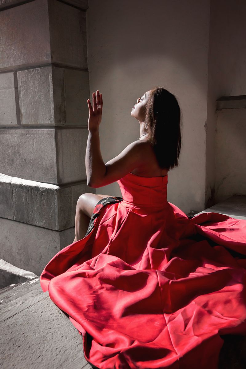 Dramatic photo of woman in flowing red dress posed against stone wall in sunlight.