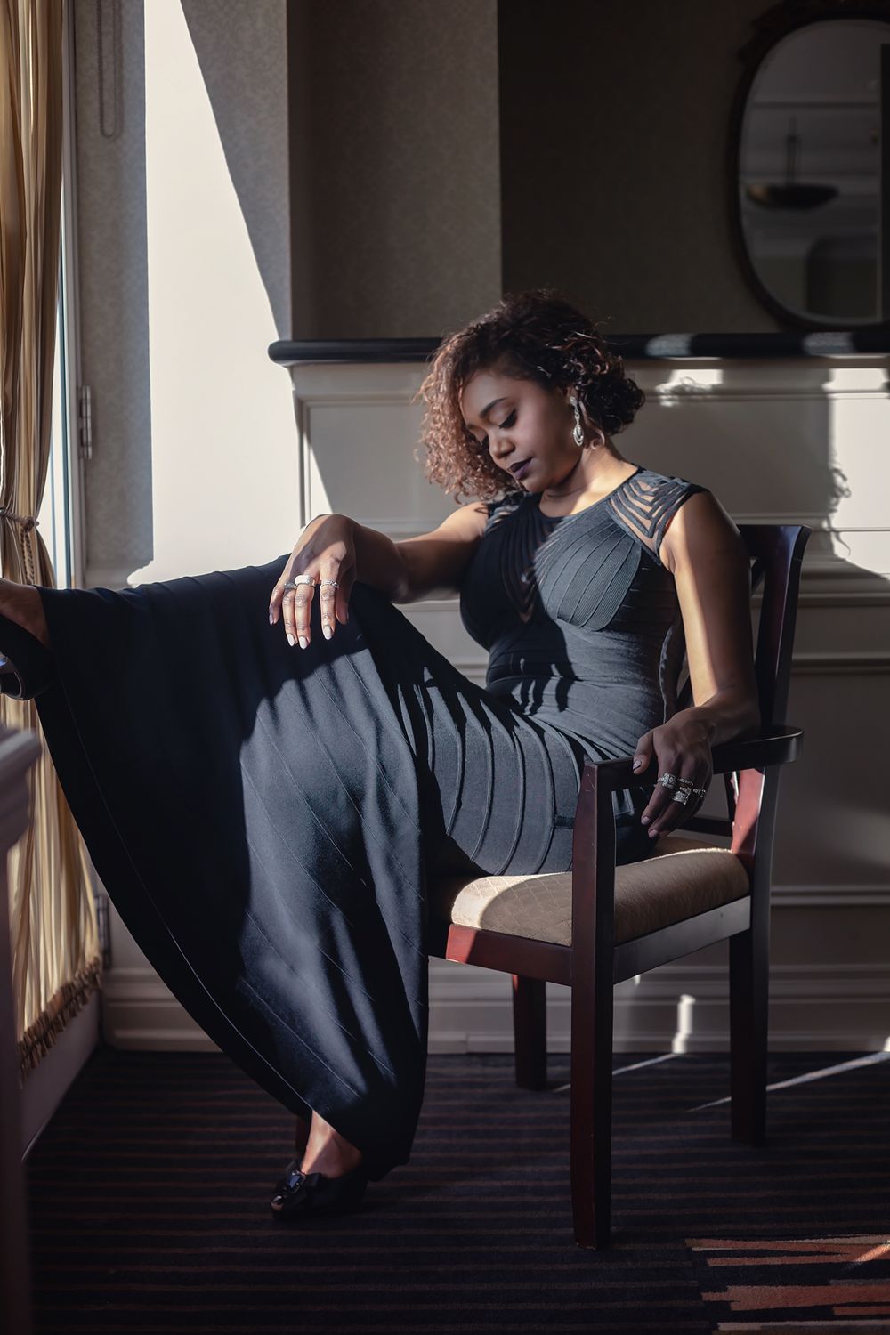 Elegant woman in navy blue dress reclining on wooden chair in moody lighting.