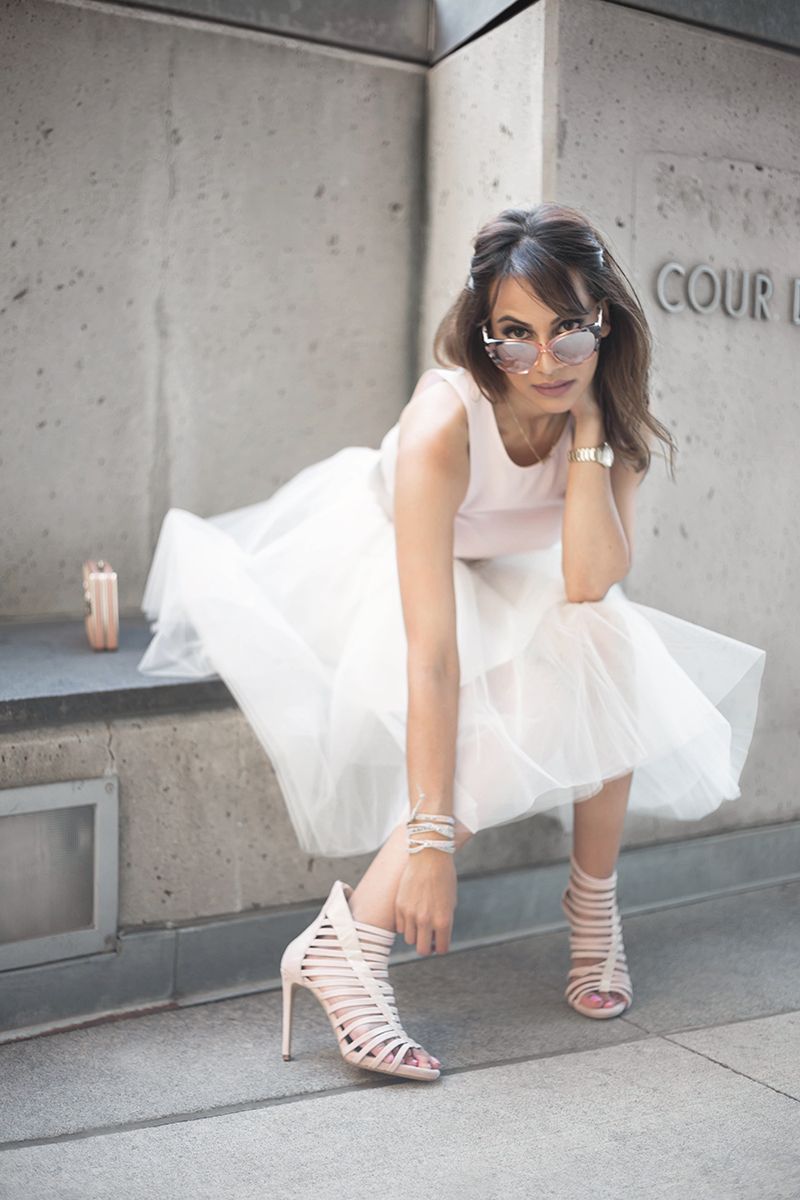 Fashion model in white tulle dress and strappy heels posing against concrete wall.