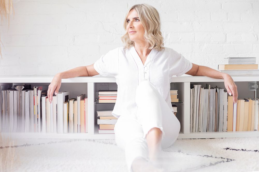 Minimalist portrait of someone in white clothing sitting against a bookshelf in a bright interior.