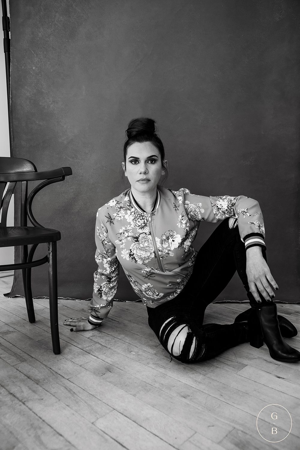 Black and white portrait of someone in a floral jacket and ripped jeans sitting on wooden flooring.