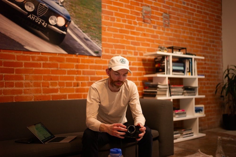 Person in casual attire sitting on couch playing video games in brick-walled room with shelving.