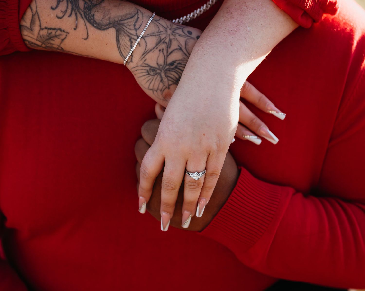 Hand with black floral tattoo sleeve and diamond ring resting on red sweater sleeve.