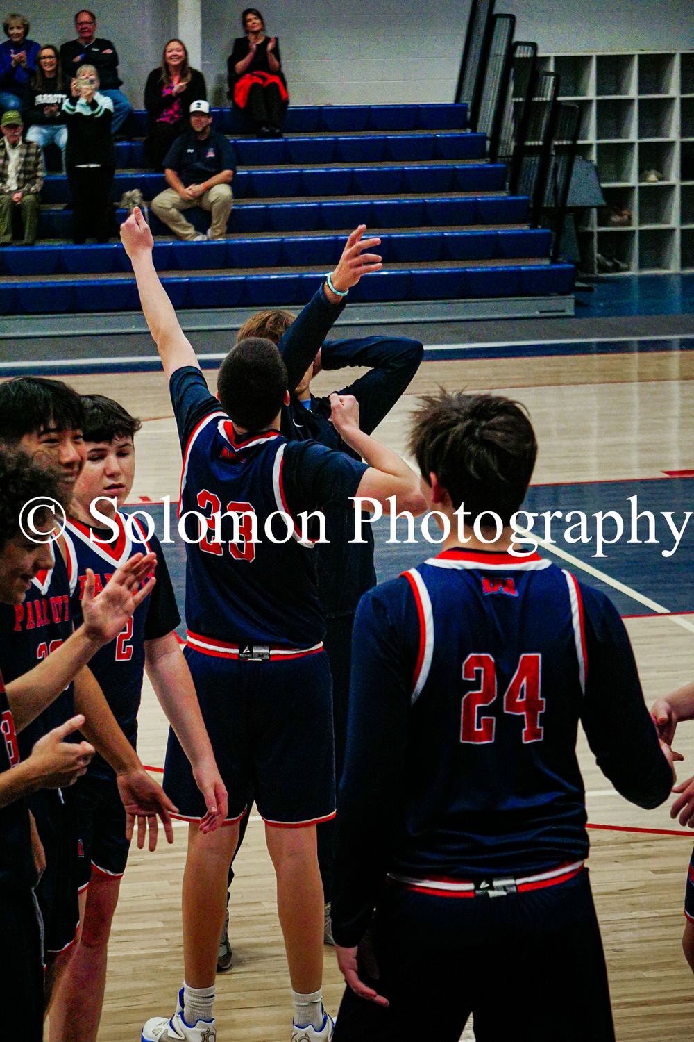 High school boys basketball team celebrates victory with raised arms on home court.