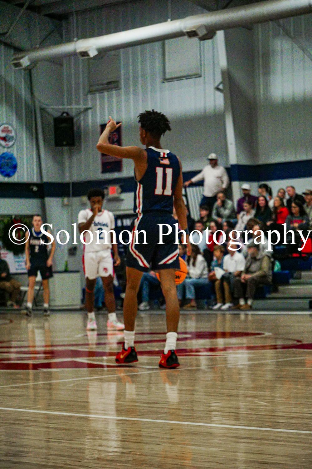 Player #11 in navy uniform signals play during high school boys basketball game.