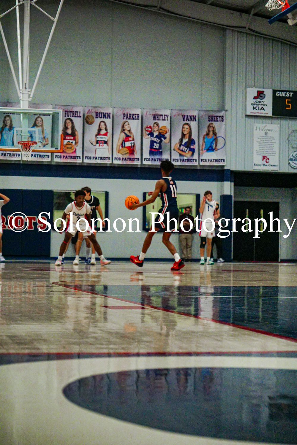 A basketball player dribbles down the court during a high school game in a gymnasium with team banners on the wall.