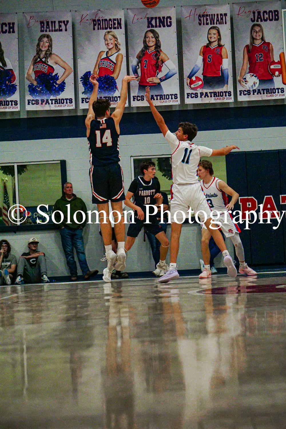 Player number 14 shoots over a defender during a high school basketball game with FCS Patriots banners in background.