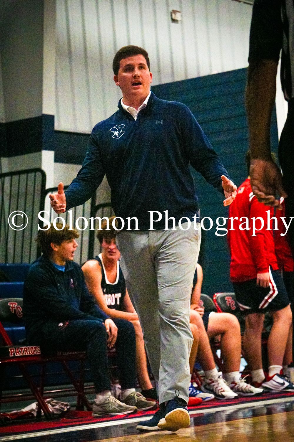 A basketball coach in a navy pullover gestures animatedly from the sideline during a high school basketball game.