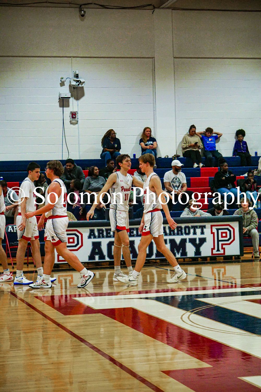High school basketball players in white uniforms gather on the court at Arena Parrott Academy gymnasium.