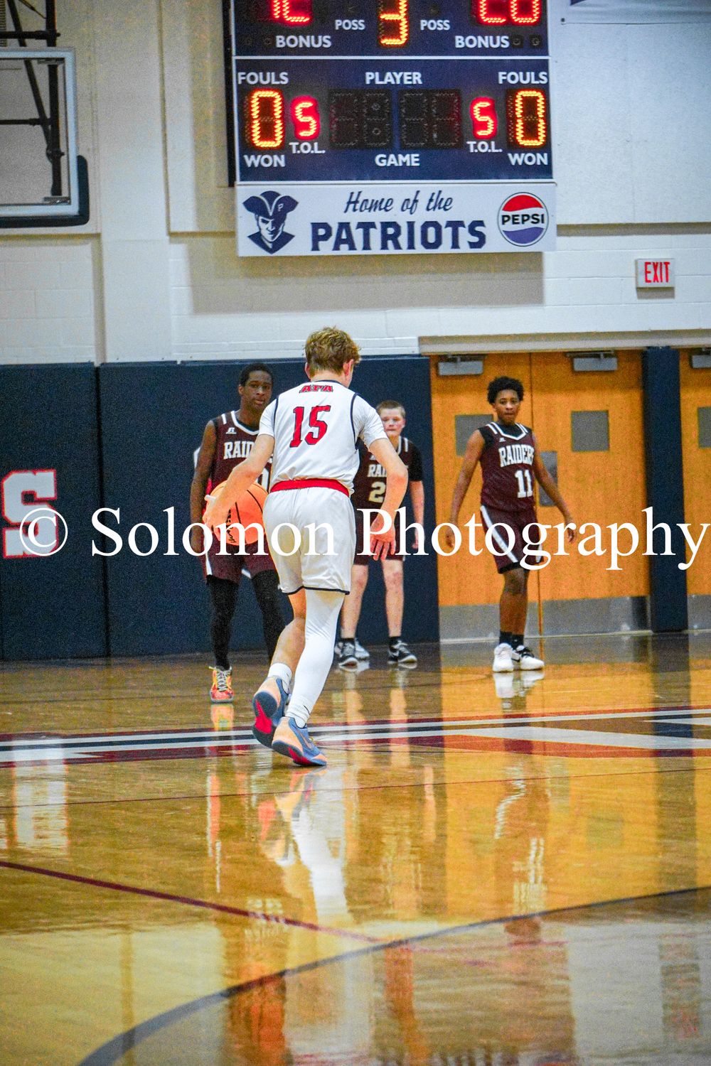 Player number 15 in white dribbles the ball near half court during a Patriots home basketball game.