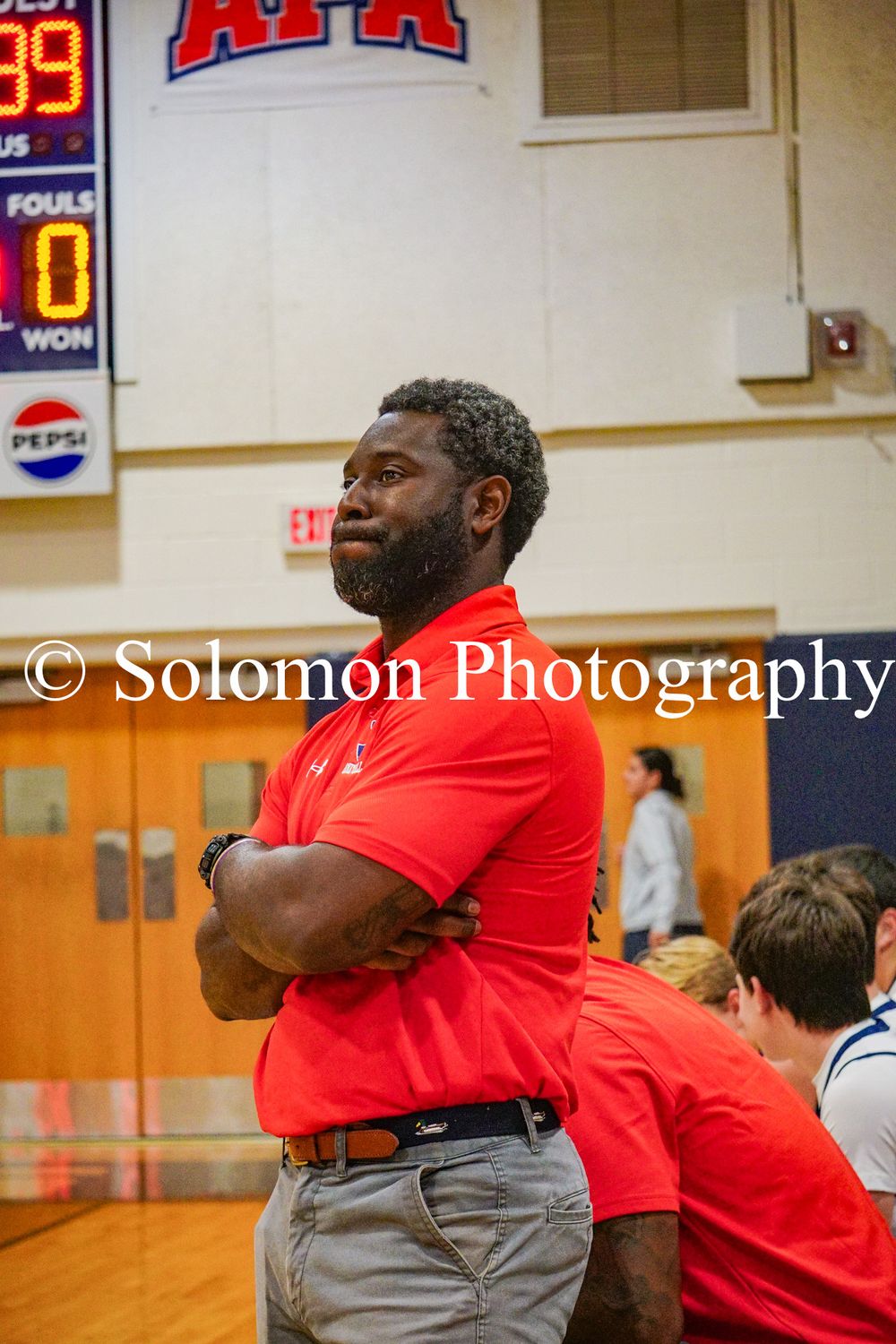 A coach in a red polo shirt stands with arms crossed watching a high school basketball game intently.