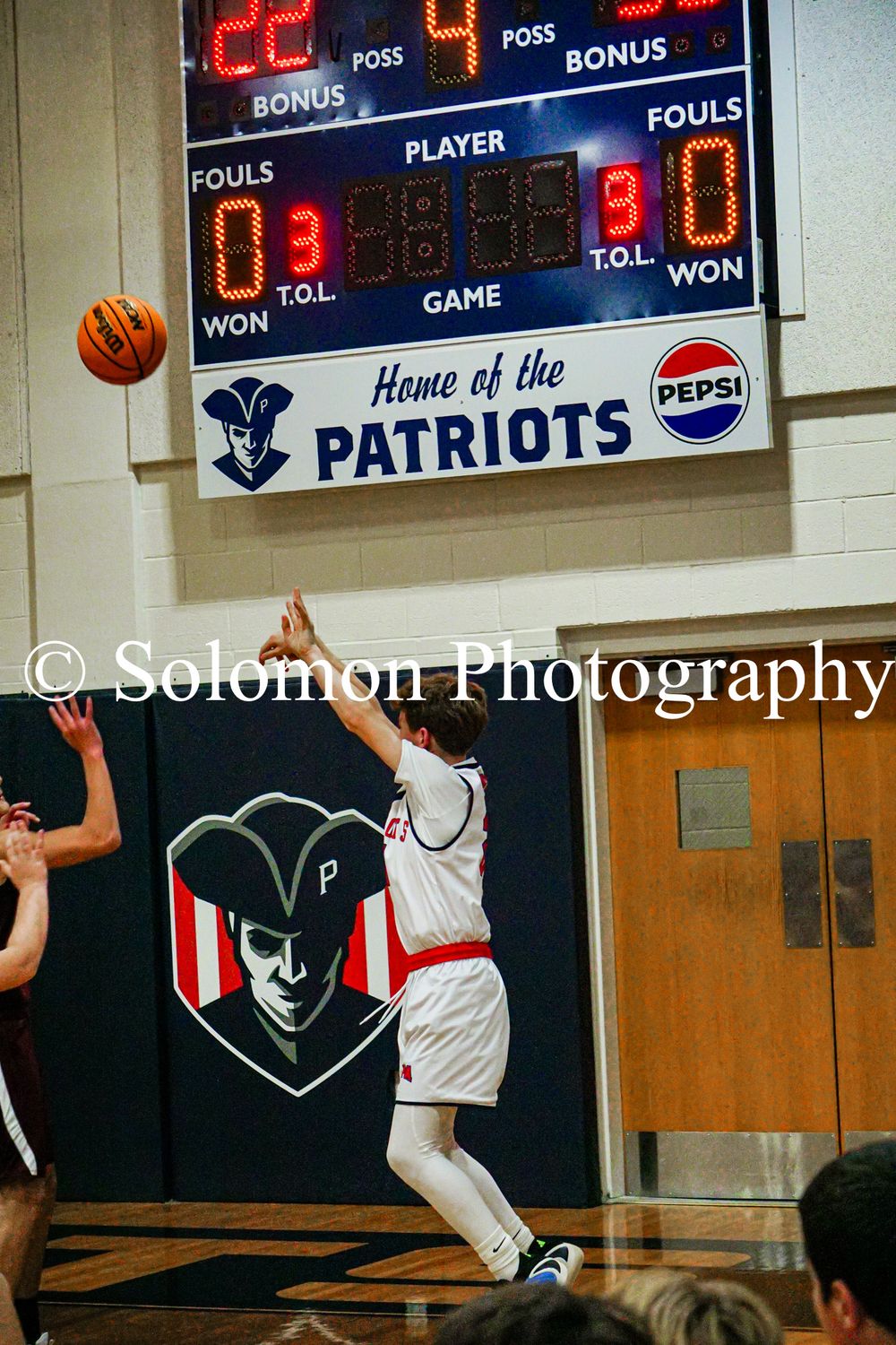 A Patriots player launches a jump shot with the scoreboard visible showing game details in the background.
