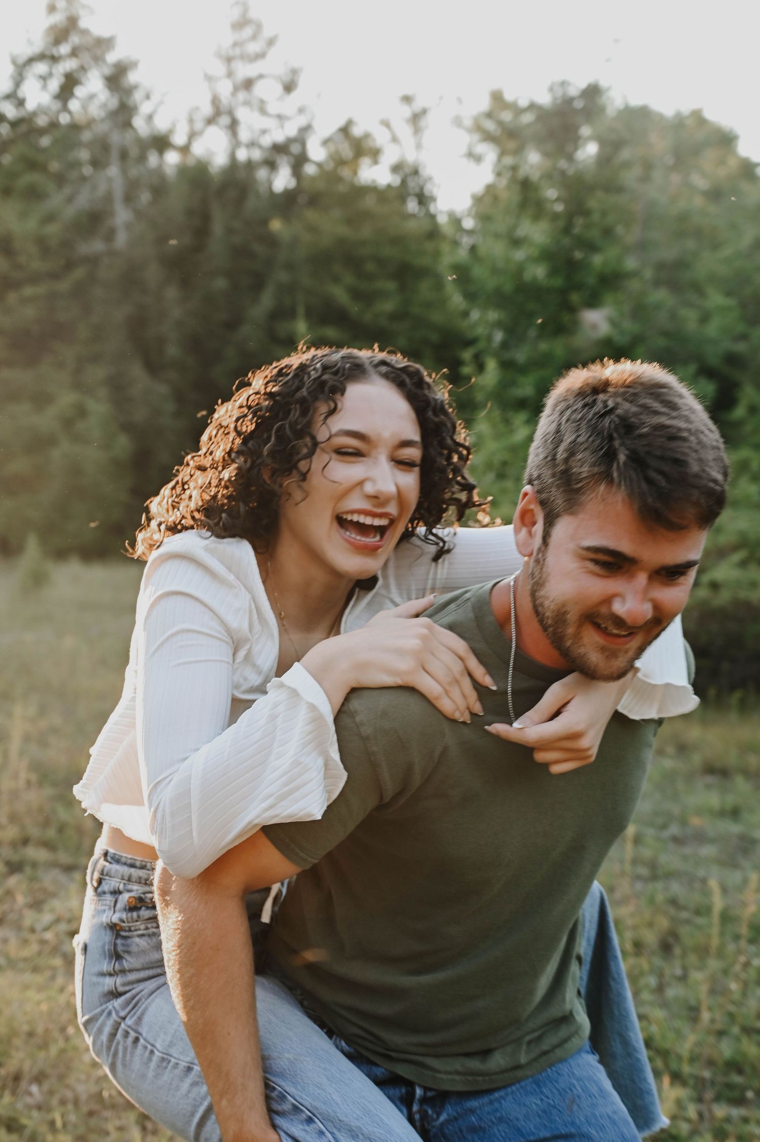 A joyful couple laughs together outdoors during a playful piggyback ride at sunset in a grassy field.