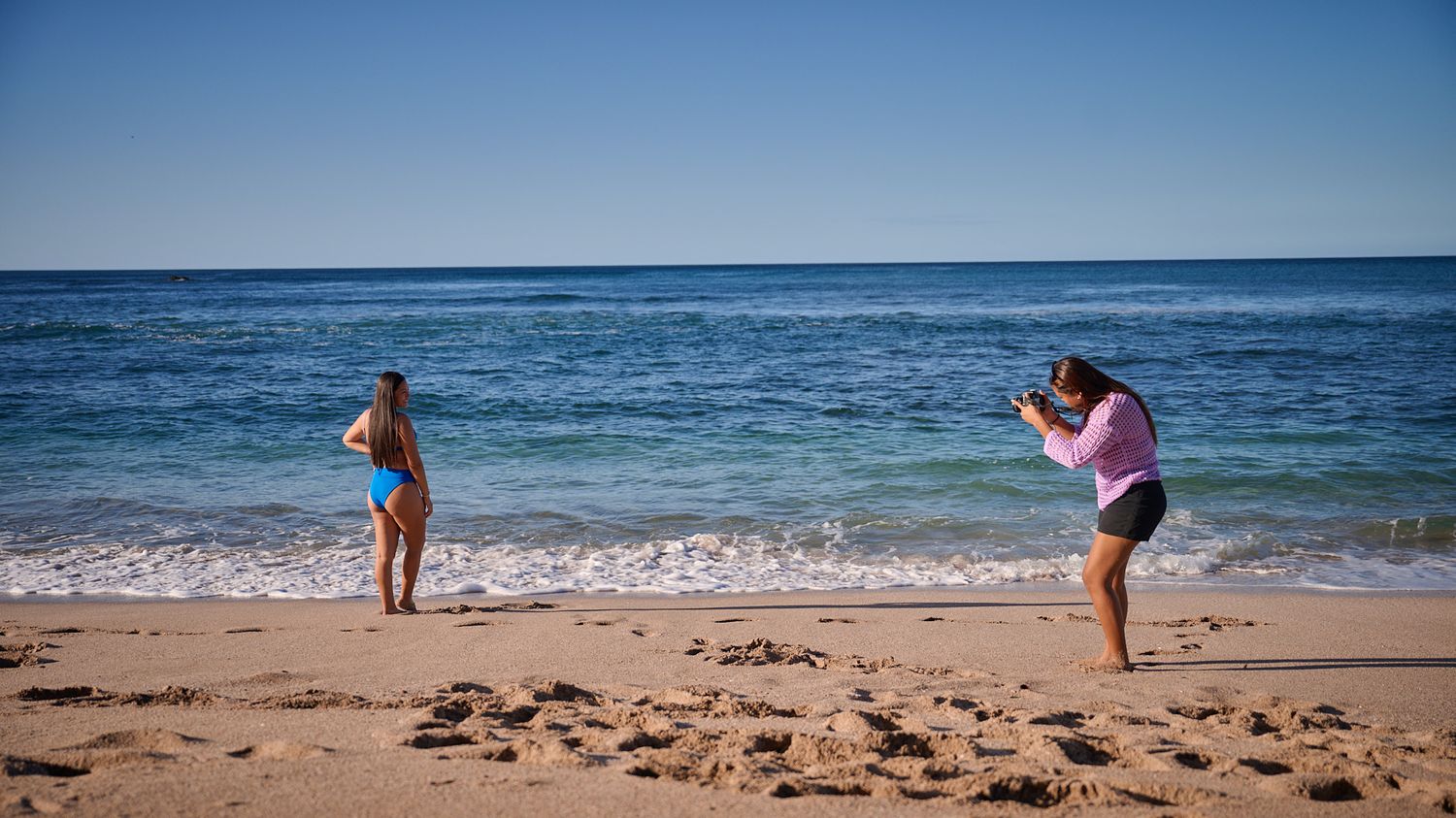 Two people on a sandy beach taking photos of each other near the ocean on a sunny day.