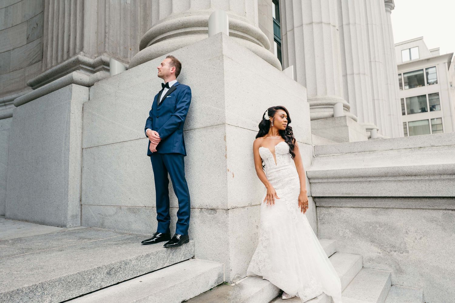Bride in white lace dress and groom in navy suit pose separately against elegant marble columns of classical building.