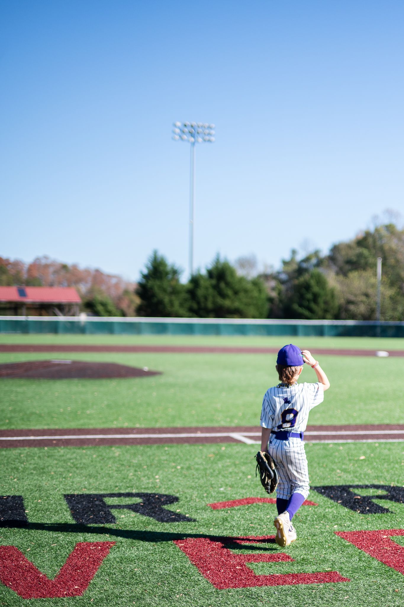 Young baseball player in blue and white uniform stands on artificial turf field with stadium lights in background.