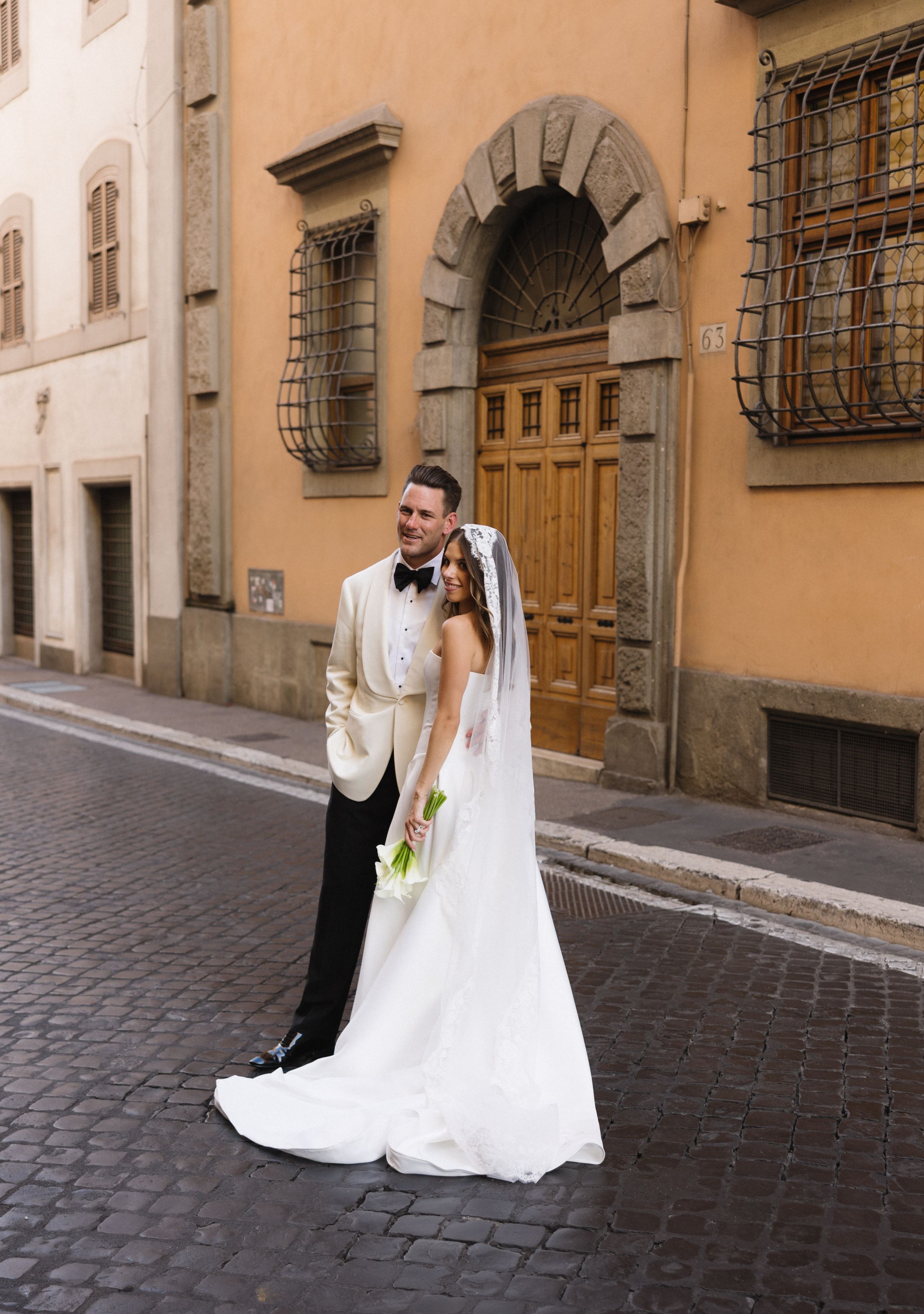 A couple in wedding attire sits together on cobblestone steps of a charming European street with historic buildings.