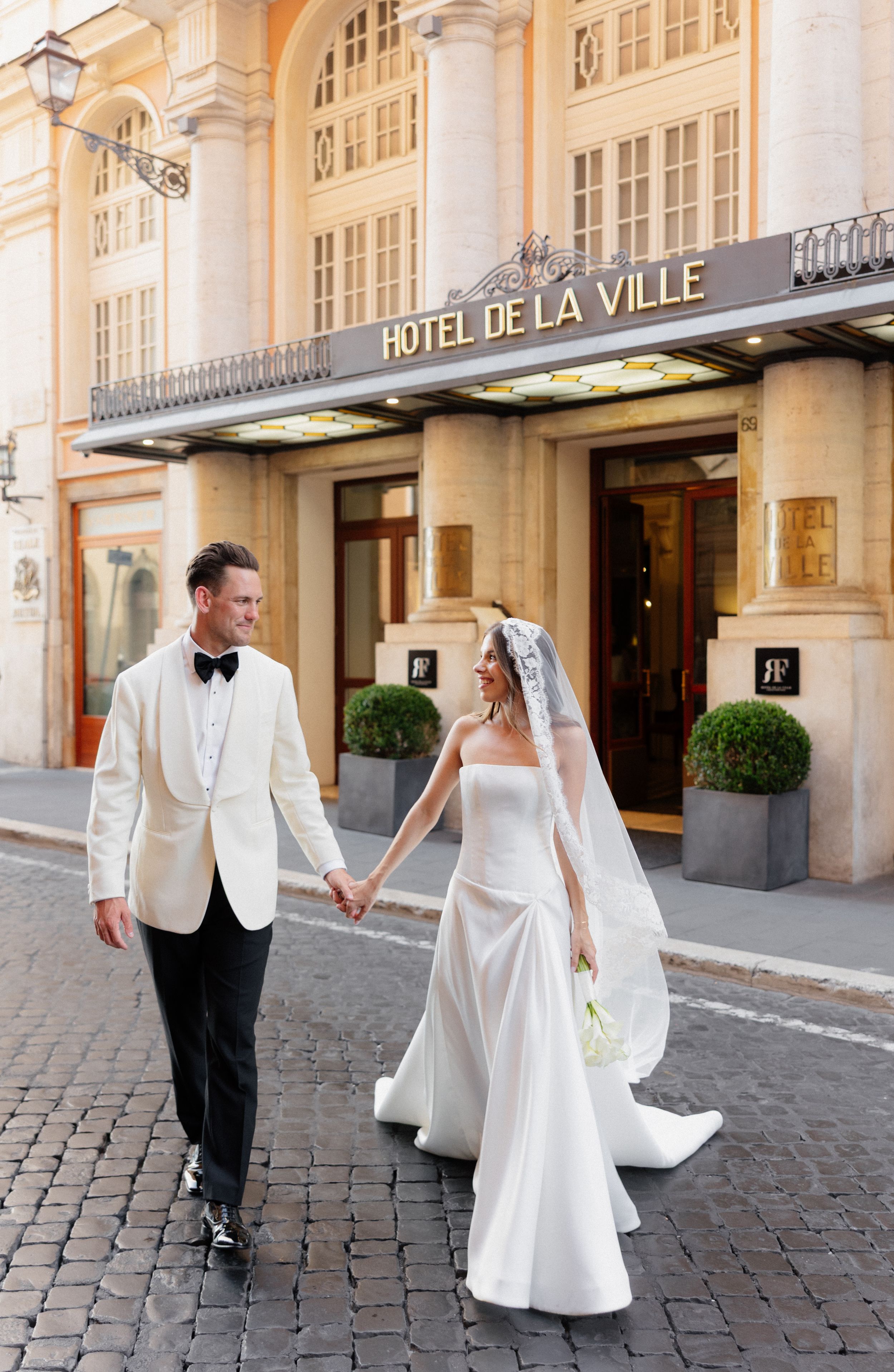 Elegant couple in white wedding attire walking on cobblestone street in front of historic Hotel de la Ville building.