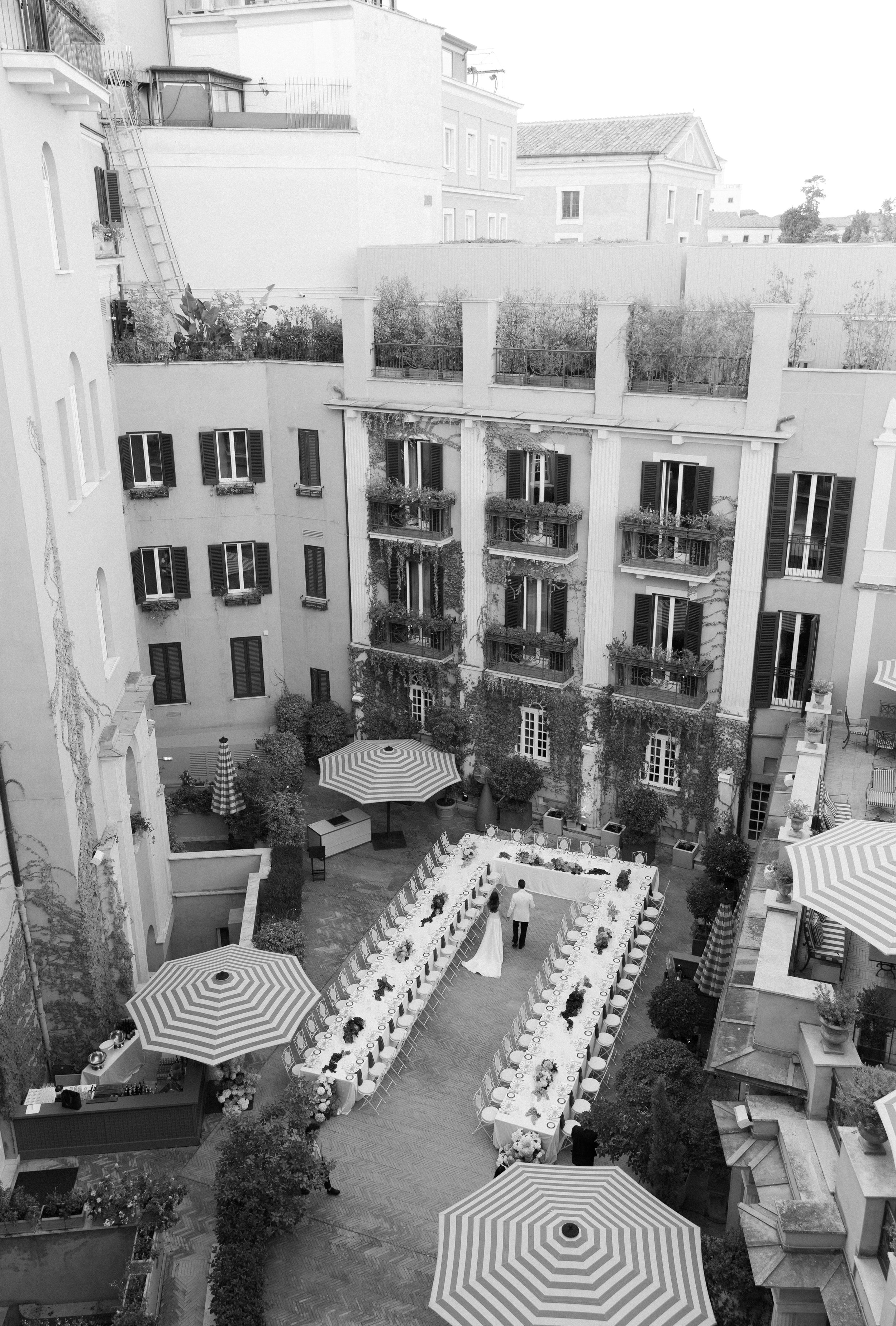 Aerial view of an elegant courtyard dinner setup with striped umbrellas and long tables arranged in a U-shape.