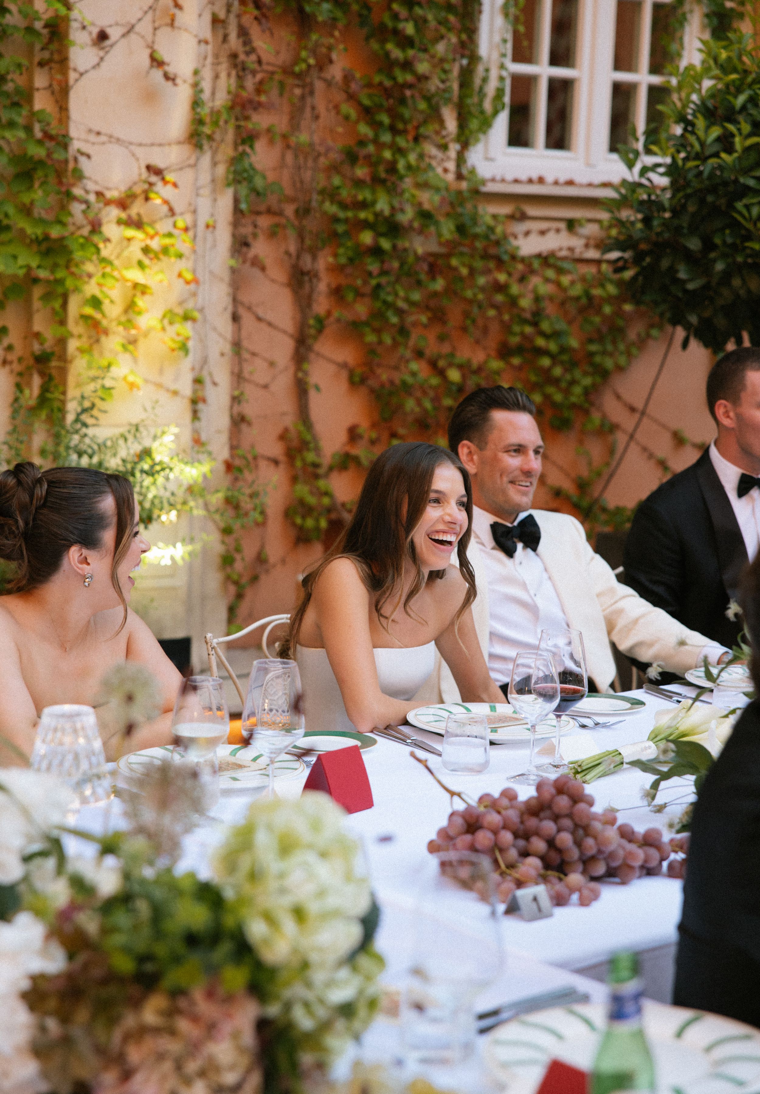 Wedding guests enjoy a joyful outdoor reception dinner at a rustic venue with ivy-covered walls and floral decorations.