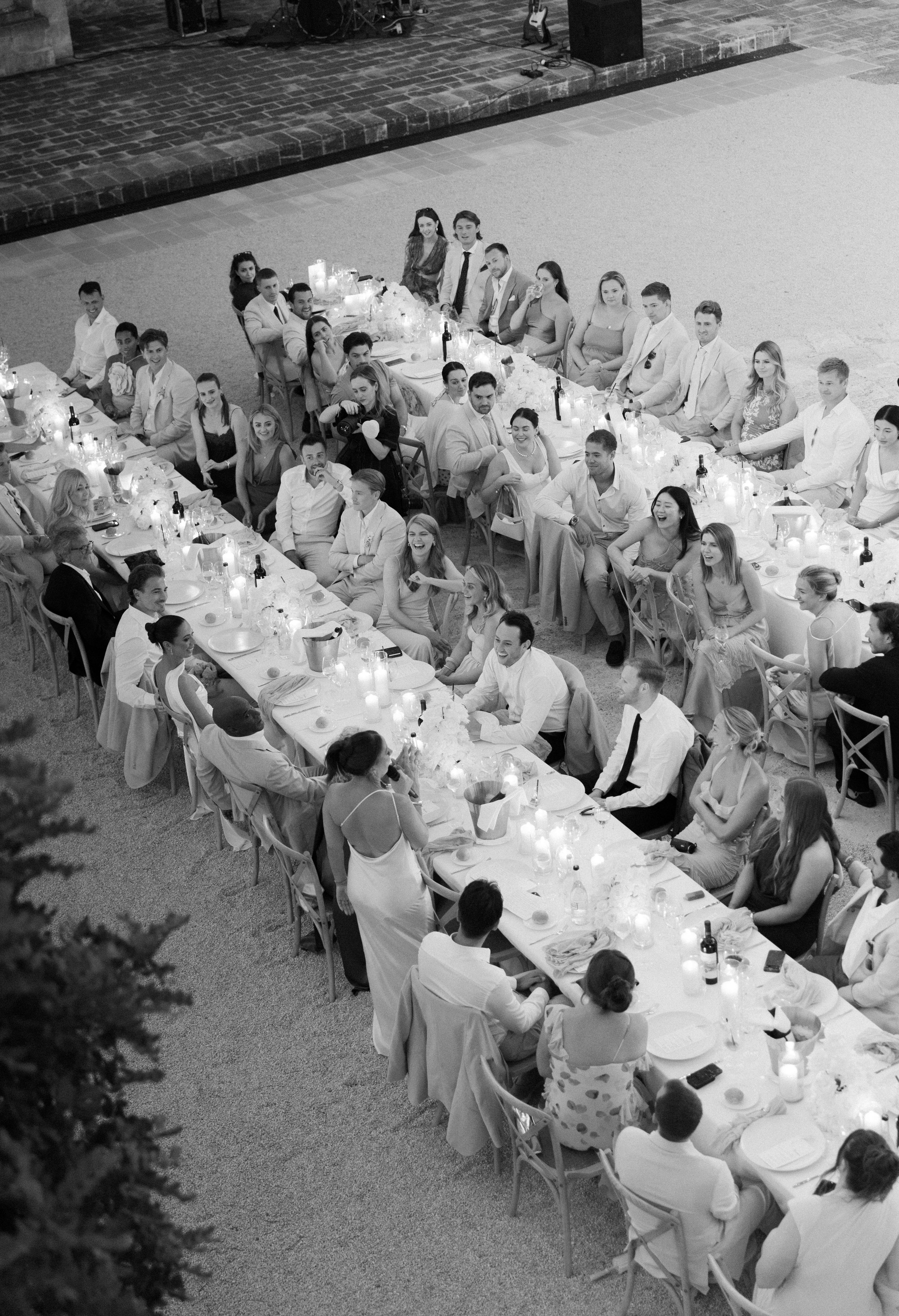 Aerial view of wedding guests seated at long tables during an outdoor reception in black and white photography.